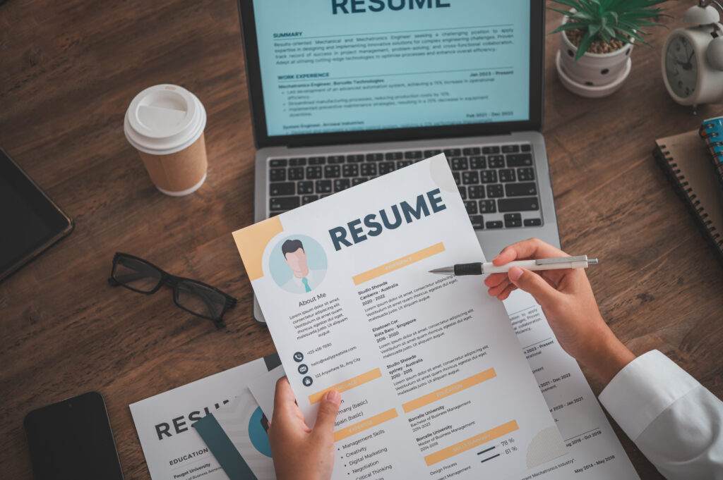 A pair of hands holding a person’s resume, with other resumes, a laptop, and a coffee cup on the table.
